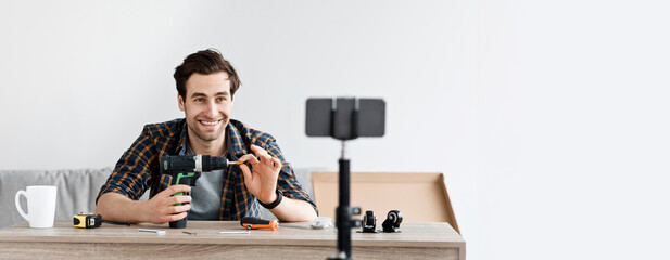 Carpenter recording video for repair blog or review of instruments. Happy attractive millennial man looks at smartphone in webcam and shows electric screwdriver, in living room interior with tools