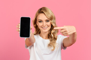 Fototapeta premium A cheerful young woman with wavy blonde hair holds up a smartphone against a vibrant pink background, pointing at it with enthusiasm and inviting attention to the device.