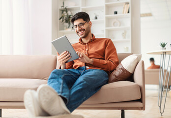 Portrait Of Excited Laughing Handsome Arabic Man Holding And Using Digital Tablet Sitting On Sofa...