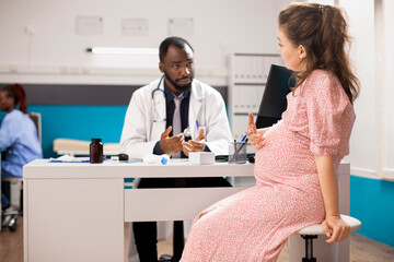 Fototapeta premium Expectant mother talks to african american obstetrician about her pregnancy progress during medical checkup. Friendly doctor listening to pregnant caucasian woman while seated at clinic office desk.