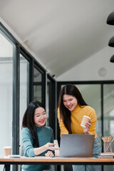 Two young women in a modern office setting, collaborating over a laptop with coffee, showcasing teamwork and creativity.