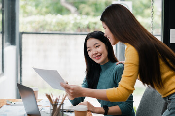 Two women working together in a bright office, reviewing documents and using a laptop, showcasing teamwork and collaboration.
