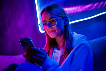 Photo of cute charming blonde girl hold device sit sofa indoors nightclub neon blue purple light...