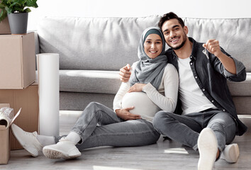 Loving expecting family handsome husband and wife in hijab with big tummy showing key from new apartment and smiling at camera, celebrating moving to own apartment, sitting on floor, copy space
