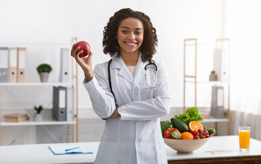 Beautiful african woman dietologist holding apple in her hand, recommending fresh fruits, posing at...
