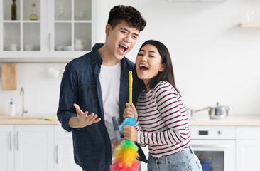 Funny asian loving couple singing songs while cleaning kitchen, using fluffy dust brush as...