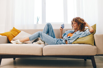 Happy woman sitting on a cozy sofa at home, holding her mobile phone and smiling while playing an online game and reading messages in cyberspace.