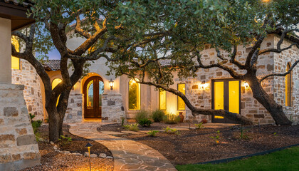 Fototapeta premium Beautiful stone house with warm lighting and lush landscaping at dusk