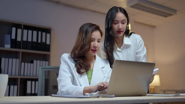 Two asian businesswomen discussing strategies while working late in an office, focused on their laptops and collaborating effectively