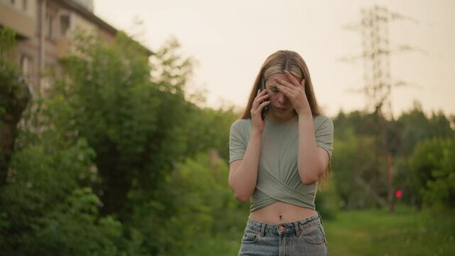 Young woman looking exhausted during phone call, with her left hand covering her forehead in frustration, surrounded by blurred greenery, with a faint green light and building in the background