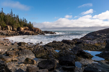 pebble beach in maine blue sky with clouds waves crashing on the rocky east coast beach pines in the background 