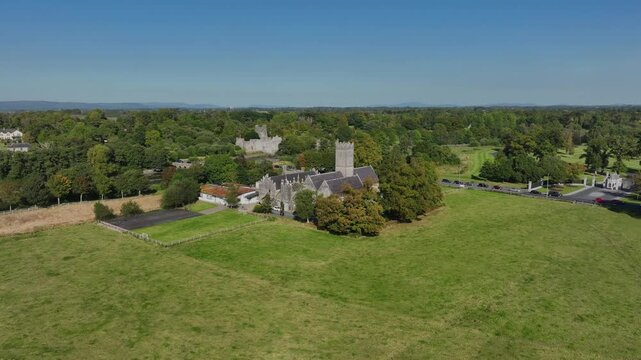 Augustinian Friary, Adare, County Limerick, Ireland, September 2024. Drone orbits counter clockwise over open grassy fields as trees obscure the historic stone building as cars drive along the road.