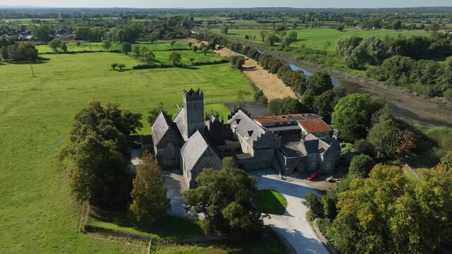 Augustinian Friary, Adare, County Limerick, Ireland, September 2024. Drone orbits counter clockwise above entrance courtyard and roundabout of church with historic grey stone facade.