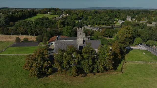 Augustinian Friary, Adare, County Limerick, Ireland, September 2024. Drone orbits clockwise revealing the gothic church building through trees as cars drive across River Maigue over a stone bridge.