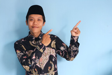 Young Asian Muslim man wearing a batik shirt and songkok pointing finger at free space place for advertisement