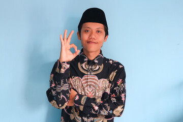 Young Asian Muslim man wearing a batik shirt and songkok showing okay sign gesture at the camera