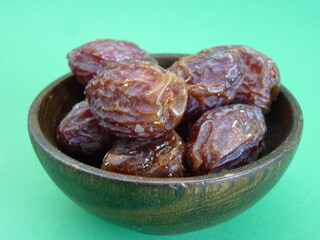 A wooden bowl filled with plump, glossy dates symbolizes abundance, nourishment, and the sweetness of life. Against a vibrant green background, these dates represent both natural simplicity.