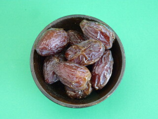 A wooden bowl filled with plump, glossy dates symbolizes abundance, nourishment, and the sweetness of life. Against a vibrant green background, these dates represent both natural simplicity.