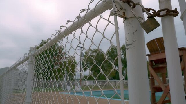 A time-lapse shot of a chain-link fence secured with a lock and chain, framing an empty outdoor pool, with overcast skies and trees in the background.