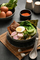 Ramen noodles served in a ceramic bowl, topped with egg, beef, narutomaki (fish cake slices), scallions (sliced green onions), and nori, placed on a bamboo mat and a black table.