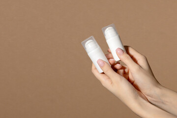 Two female hands are holding White unmarked bottles for cosmetics. Mock up of container for serum, lotion, cream, retinol. Brown background with copy space