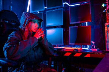 Obraz premium Woman hacker. A woman wearing a hood is thinking looking at a computer monitor. The room is dimly lit. possibly typing or browsing the internet, Hacker in neon light, Female gamer think