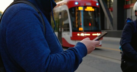 Man holding smartphone while waiting for tram in North American city. Scene showcases modern mobility solutions and reliance on technology for seamless urban commuting experience.