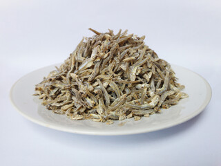 A pile of dried anchovies, a popular Indonesian snack called Ikan Teri, on a white plate. The fish are small, silvery, and have a slightly curved shape. They are a good source of protein and calcium.
