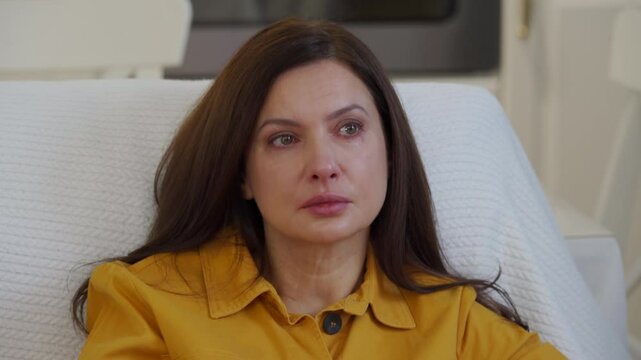 Adult 44 year old woman sitting on sofa in living room in front of TV watching melodrama. Impressionable woman cries wiping tears worried while watching movie.