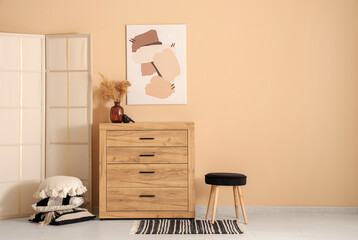 Chest of drawers, pouf and pillows near beige wall in room