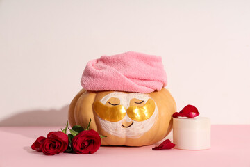 Funny pumpkin with under-eye patches, cosmetic product and rose flowers on color table against light background