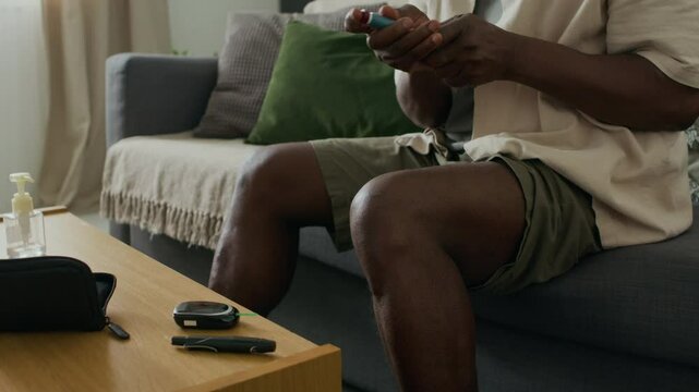 Midsection shot of African American man administering insulin injection into leg using pen device while sitting on sofa near table with diabetes supplies