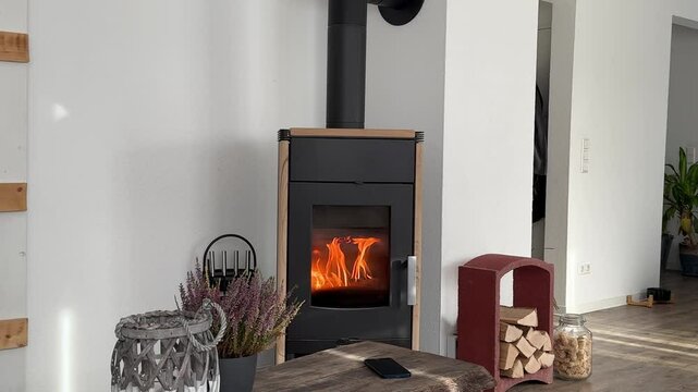A cozy interior featuring a modern black wood-burning stove with visible flames, a smartphone resting on a rustic wooden table, firewood storage, and decorative accents like a wicker lantern.