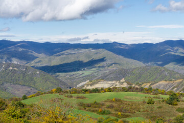 Naklejka premium Green fields, autumn bushes illuminated by the sun. Mountains, hills and sky with clouds in the background