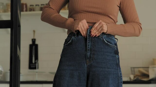 Close-up of overweight attractive mature woman trying to zip small pants after gaining weight while standing in domestic room. Midsection.