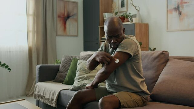 Middle-aged African American man sitting on sofa in living room, applying continuous glucose meter sensor to his arm to monitor blood sugar level