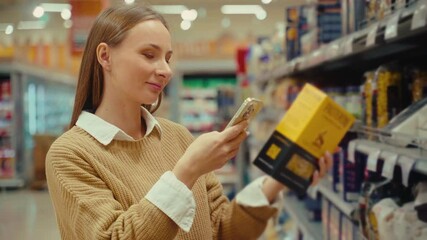 A woman uses her smartphone to scan grocery items while shopping in a supermarket aisle. She is focused on finding the right products among the shelves filled with various goods.
