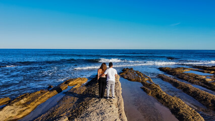 A couple stands on rocky shores, gazing at the vast ocean under a clear blue sky. The scene evokes romance and tranquility, perfect for celebrating Valentine's Day in a serene, natural setting.