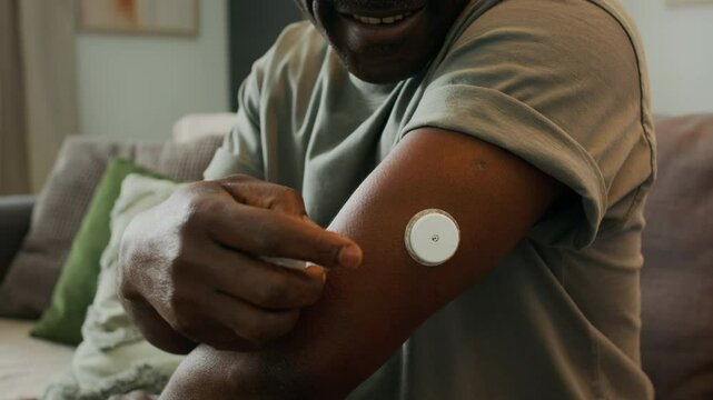 Close up of Black man with diabetes applying continuous glucose meter sensor to his upper arm while sitting at home