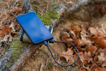 A portable charger charges the smartphone. Power Bank with cable in the forest.