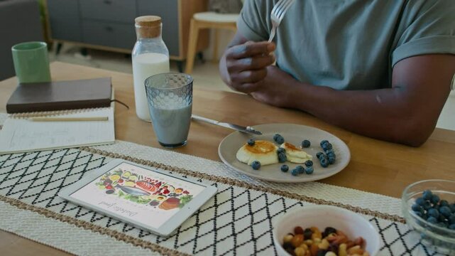 Close up view of unrecognizable Black man with CGM sensor on arm eating quark pancakes with blueberries for breakfast, digital tablet displaying healthy food on kitchen table next to him
