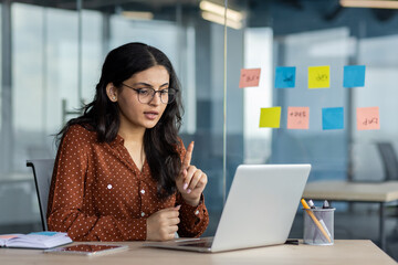 Hispanic woman businesswoman participating in virtual meeting on laptop. Displays professional communication, focus, and remote work in modern office. Ideal for business and technology concepts.