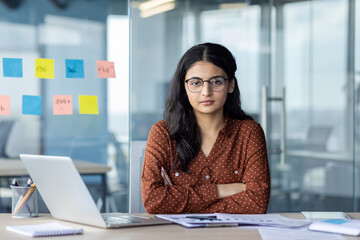 Hispanic woman businesswoman at desk with crossed arms and laptop in modern office. Scene captures determination, professionalism, focus amidst glass walls and colorful sticky notes.