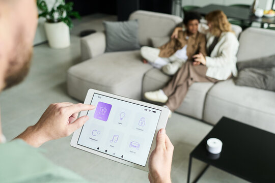 Individual using tablet for controlling smart home features while couple sits on sofa in background. Tablet displays various application icons for home automation