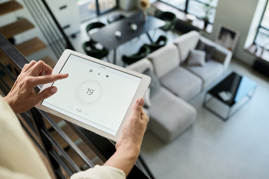 Closeup of person holding tablet and adjusting smart home temperature settings from balcony. Living room furniture and decorations visible in background