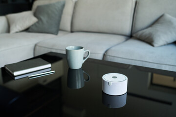 Smart speaker placed on glass table with notebook, pen, and mug, set in modern living room with beige sofa and pillows, creating a cozy and convenient atmosphere