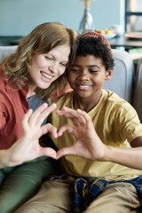 Obraz premium Mother and son forming heart shape gesture with hands while sitting on couch, sharing bonding moment and happiness. Caucasian mother and Black child smiling warmly