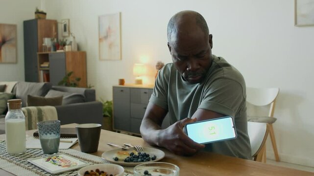 Senior African American man with diabetes checking glucose levels with smartphone and arm sensor after eating low-glycemic breakfast of blueberries and pancake in kitchen at home