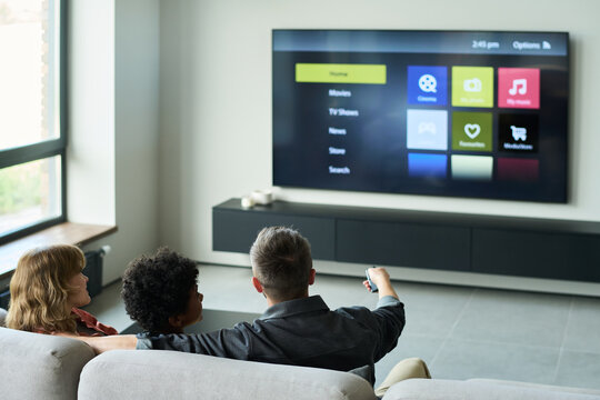 Group of people sitting on a couch watching TV. TV menu displaying various options with one person holding a remote control while others are engaged in watching screen