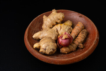 Indonesian traditonal spices for cooking, consisting of ginger, red onion, and turmeric, placed on a rustic clay plate, with solid black background